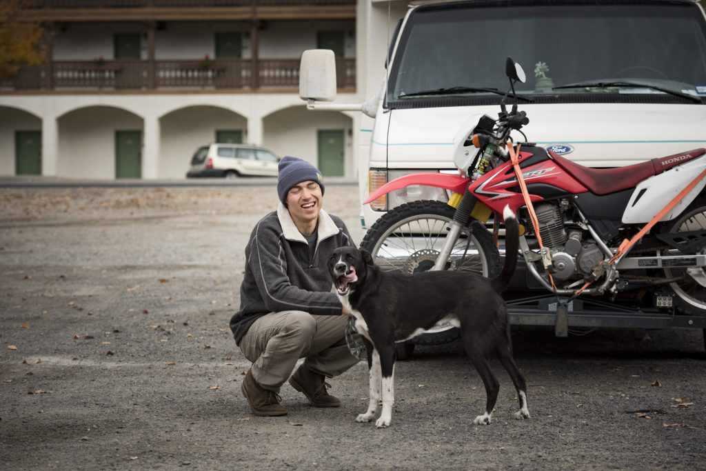 A man clothed in KUHL Men's clothing crouching next to his van with his dog.