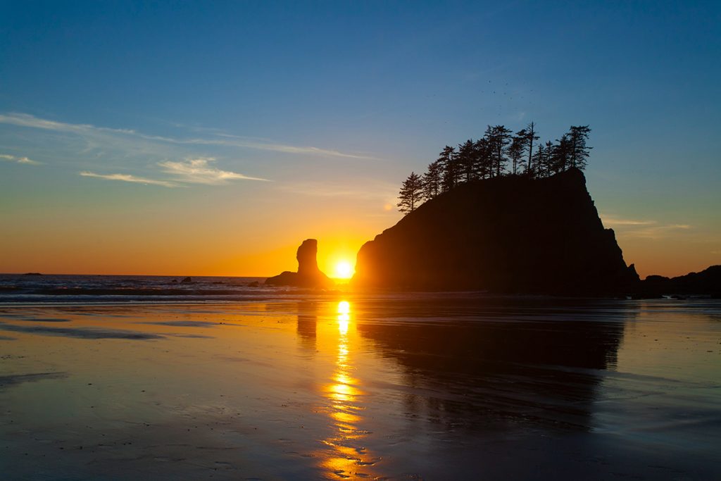 rocky structure with trees on top rising from water with sunset in background