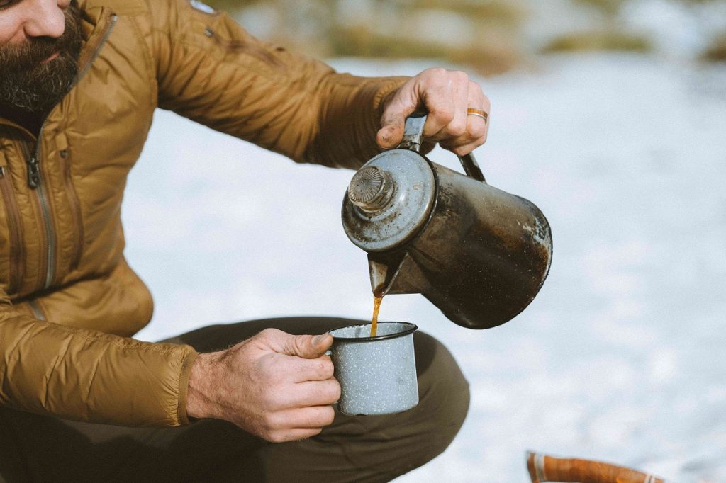 man in yellow jacket holding pot and mug on snowy ground