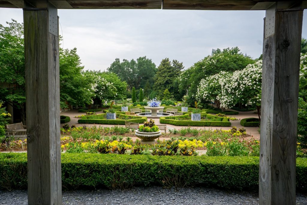 white fountain in middle of green plants trees and colorful flowers