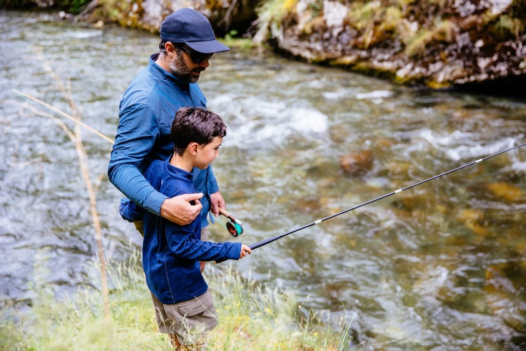 man in KÜHL shirt and KÜHL hat fishing with his son next to river