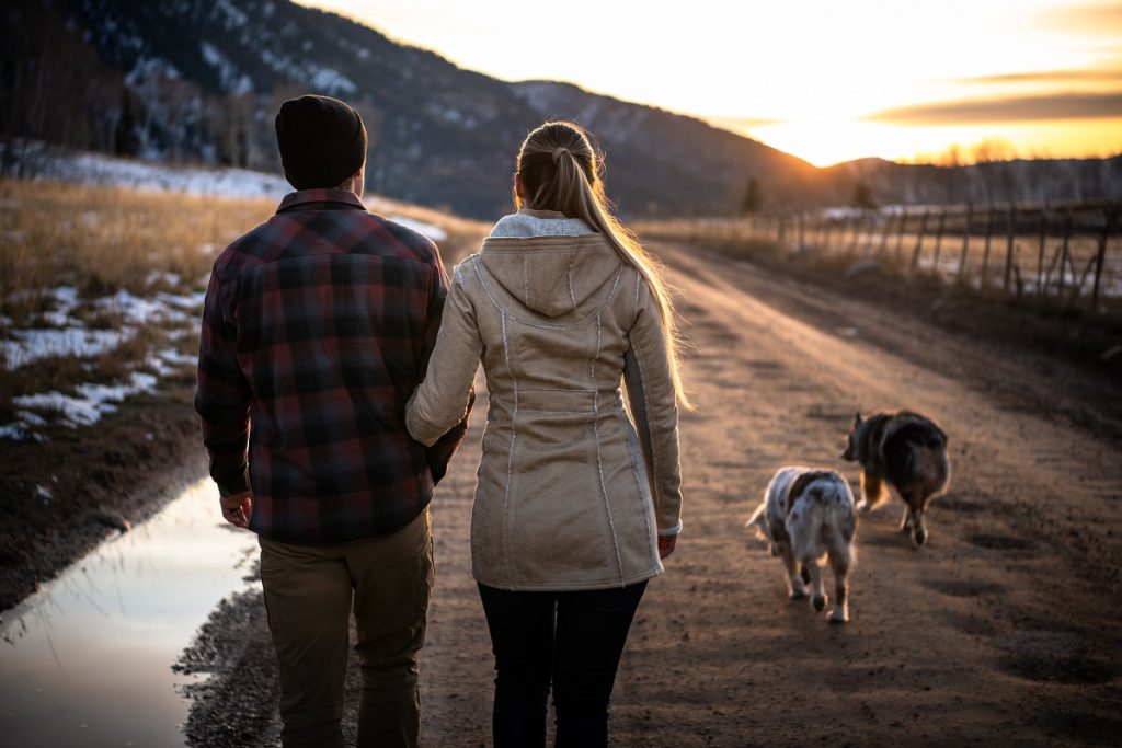 man in KÜHL plaid shirt and woman in KÜHL trench walking outdoors with two dogs